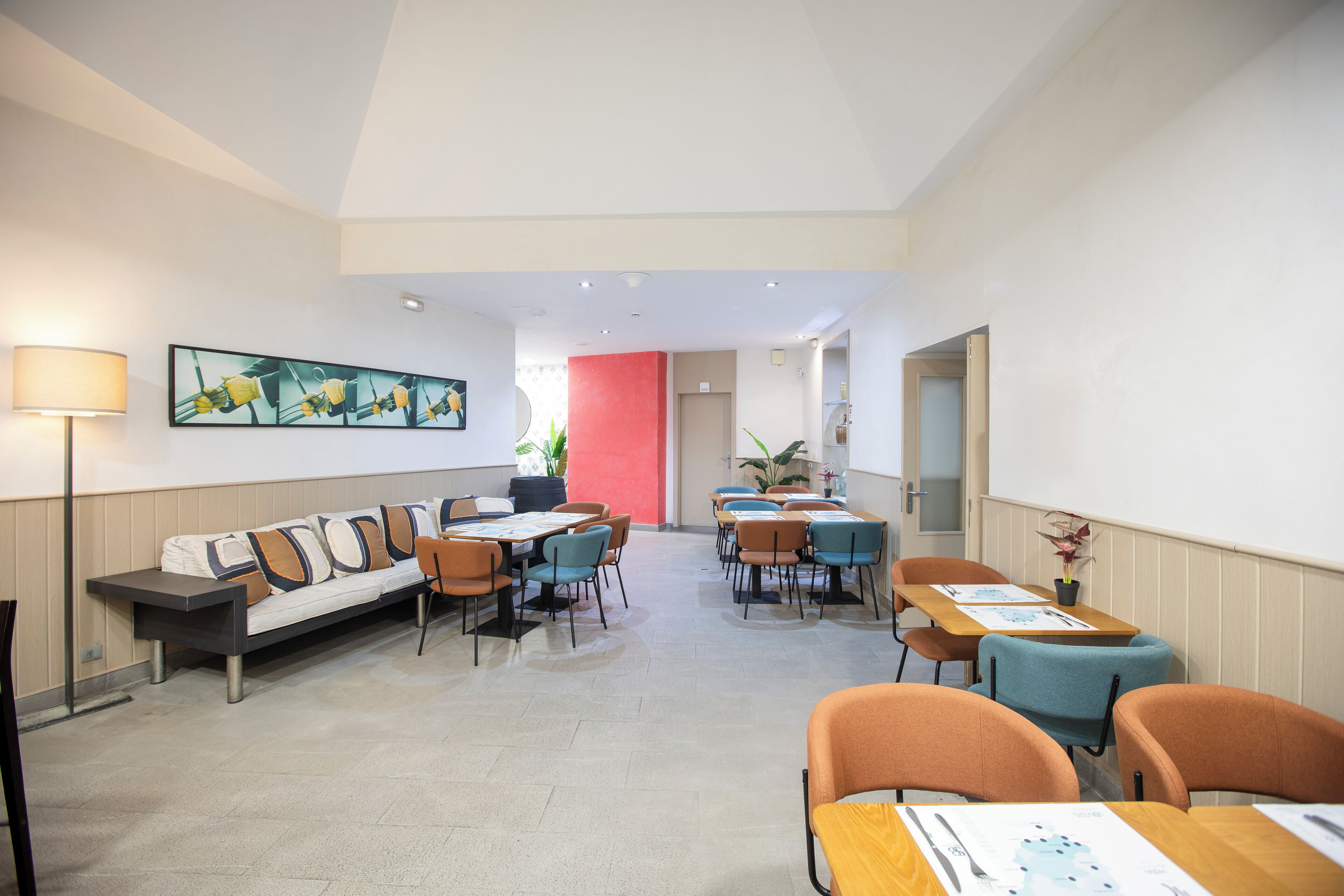 Un moderno vestíbulo de hotel con un sofá largo, mesas de comedor y sillas, un cuadro grande en la pared, y una pared de acento rojo brillante.