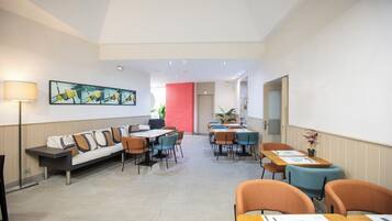 Un moderno vestíbulo de hotel con un sofá largo, mesas de comedor y sillas, un cuadro grande en la pared, y una pared de acento rojo brillante.