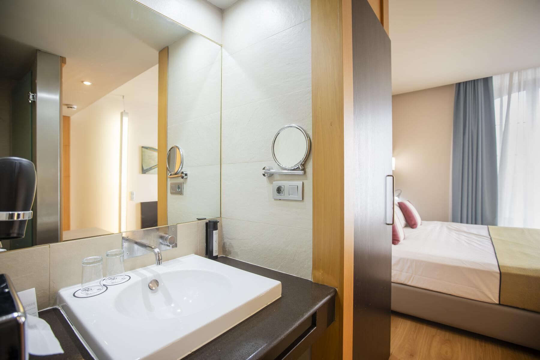 Habitación de hotel con un espejo grande, un lavamanos blanco, una cama y vistas al exterior a través de una ventana.