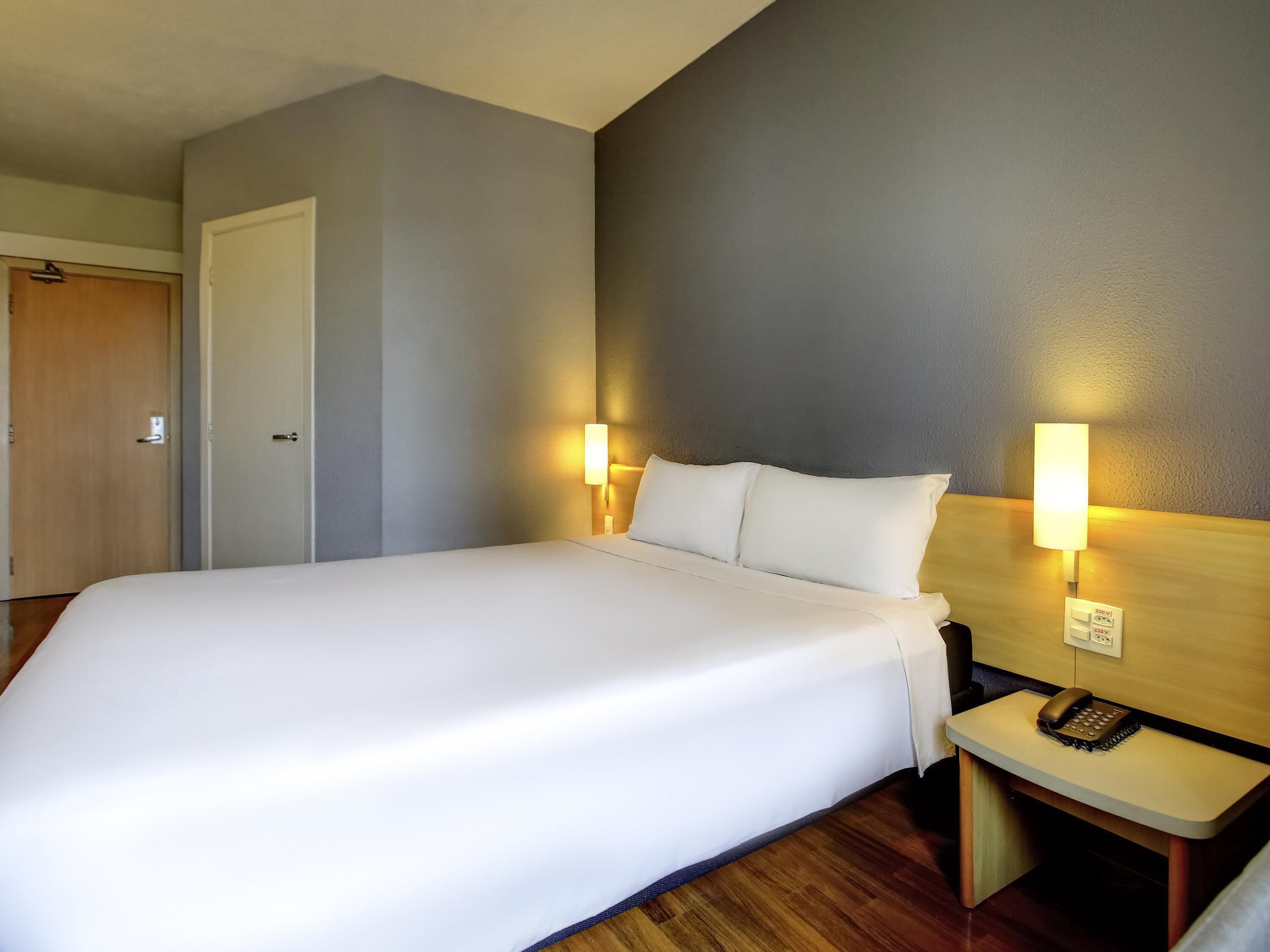Habitación de hotel con una cama grande, dos lámparas de noche, una mesita de noche de madera con un teléfono y una puerta a la izquierda.