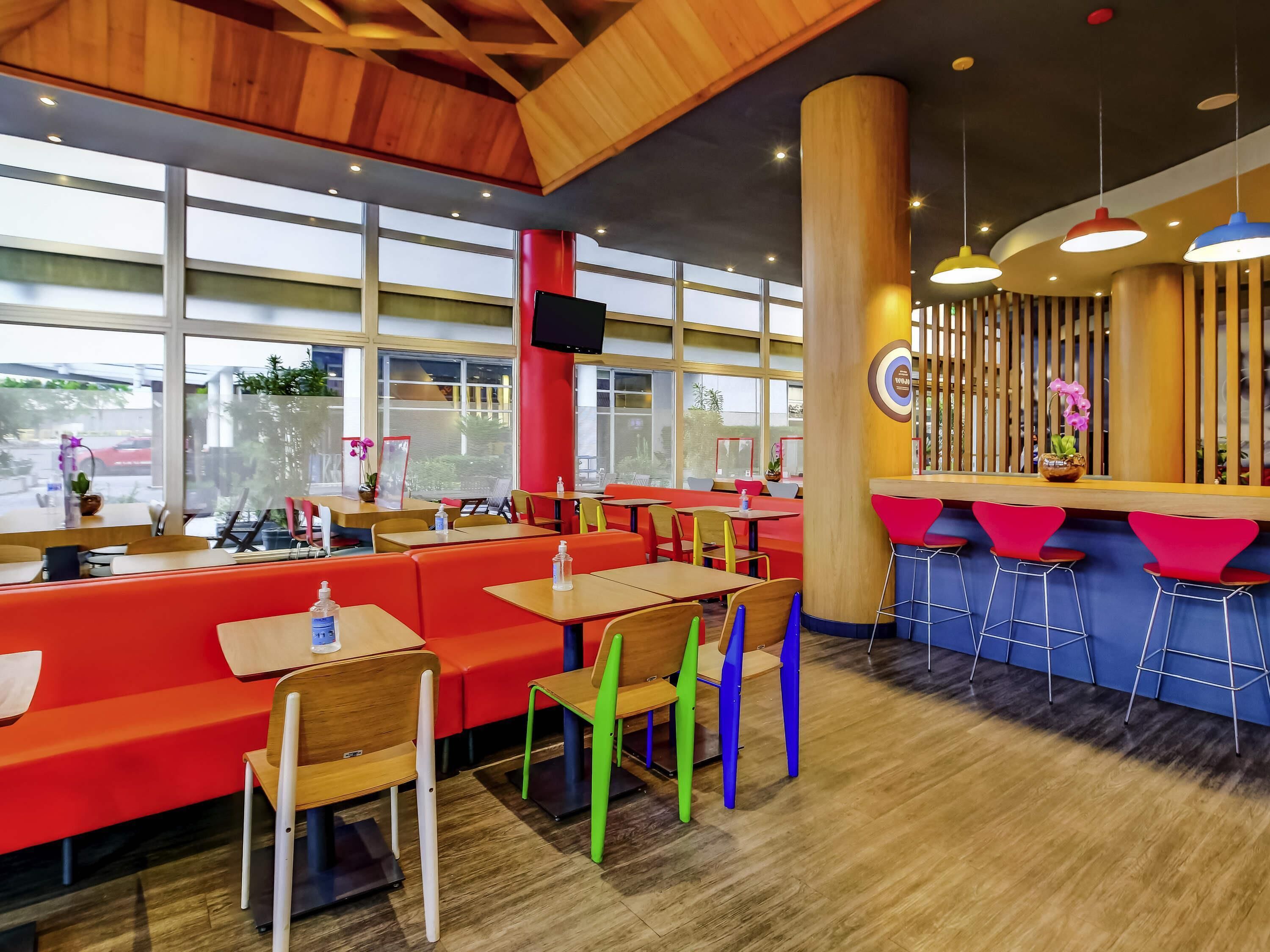A restaurant interior with wooden ceiling beams, large windows, red seating, and colorful chairs.