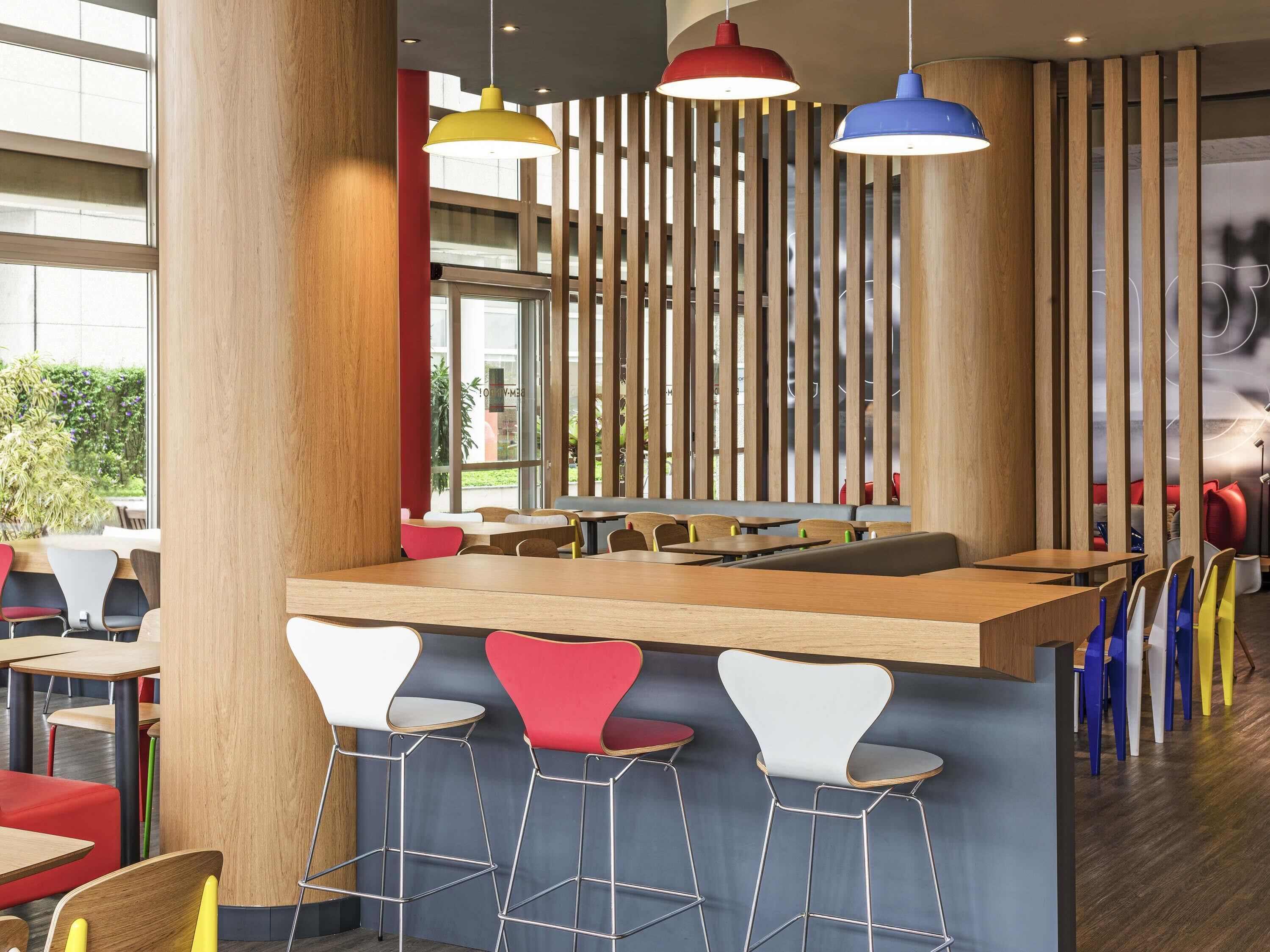 A modern restaurant interior with high wooden columns, a central bar area, and colorful pendant lighting.