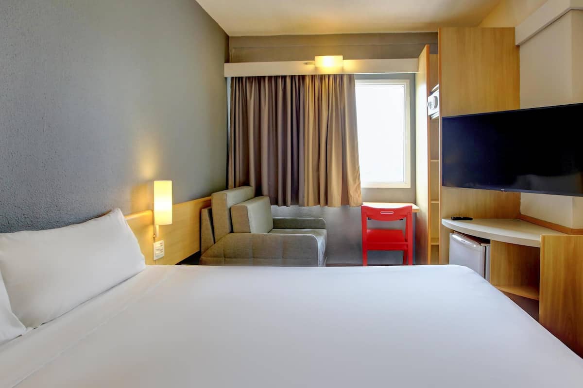 A hotel room with a large bed, a desk with a flat-screen TV, a small table, a chair, and a window with curtains.
