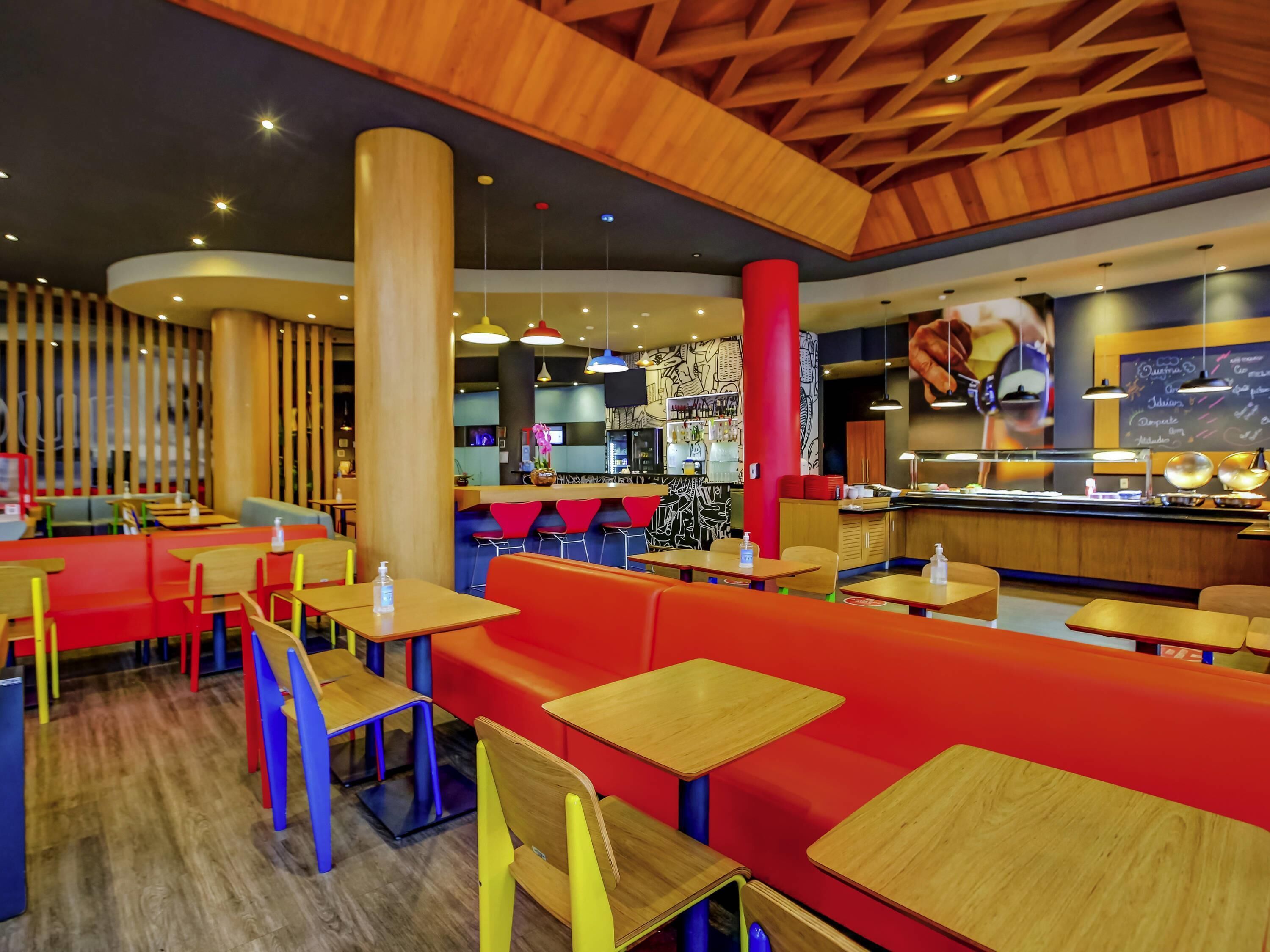 L’intérieur d’un restaurant est agrémenté de poutres en bois au plafond, de sièges rouges et bleus, et d’un bar où le menu est écrit sur un tableau noir.
