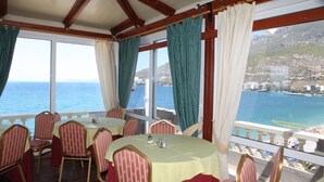 A dining area with tables and chairs, large windows with curtains, and a view of the sea and a mountainous coastline. - Mantas Seaside Boutique Hotel (Loutraki-Agioi Theodoroi)