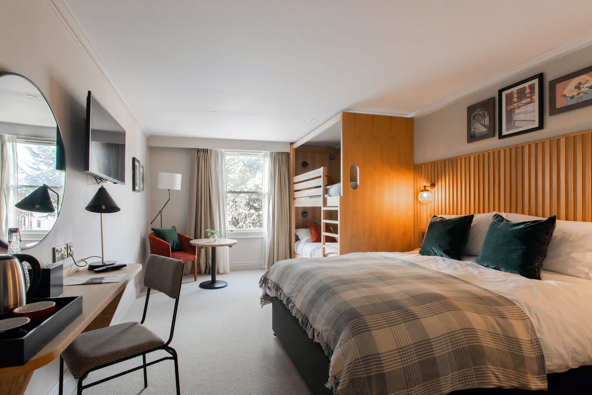 A hotel room with a bed, a desk with a chair, a TV, a window with curtains, and a wooden panel wall.