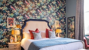 A bedroom with a floral wallpaper, a large bed with multiple pillows, bedside tables with lamps, and a nightstand with a clock.