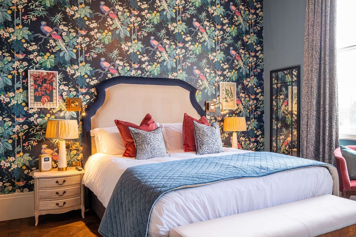A bedroom with a floral wallpaper, a large bed with multiple pillows, bedside tables with lamps, and a nightstand with a clock.