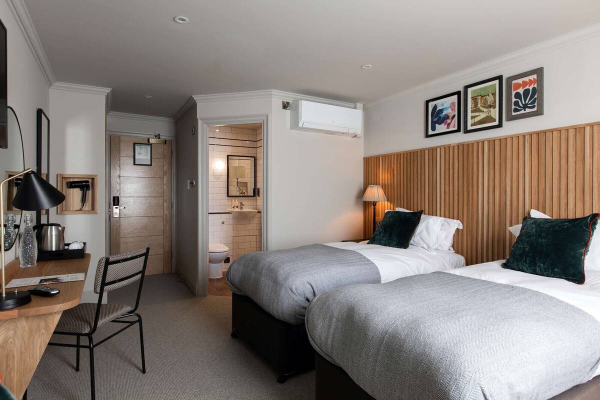 A hotel room with two beds, a desk with a chair, a mirror, and a wooden headboard.