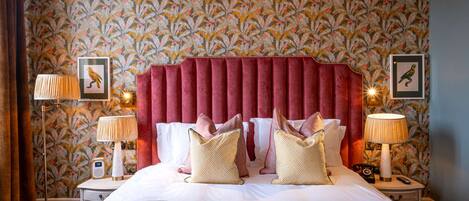 A hotel room with a large bed, red headboard, floral wallpaper, two bedside lamps, and framed pictures on the wall.