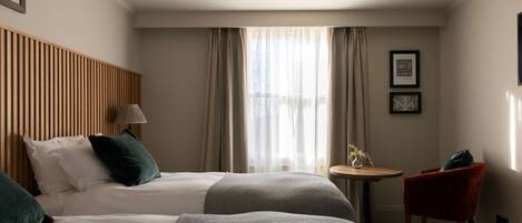 A hotel room with two beds, a small table, a chair, a lamp, and a window with curtains.