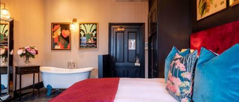 Ein Hotelzimmer mit einem großen Bett, einer roten gesteppten Bettdecke, einer weißen Wand, einer schwarzen Tür, einer weißen Badewanne und einem Spiegel mit einer Blumenvase.