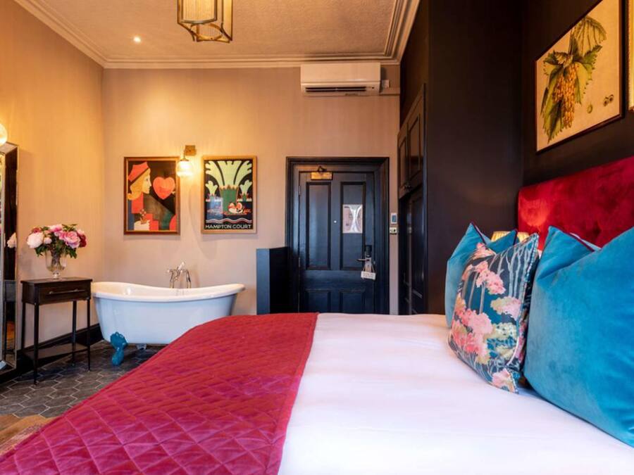 A hotel room with a large bed, a red quilted bedspread, a white wall, a black door, a white bathtub, and a mirror with a vase of flowers.
