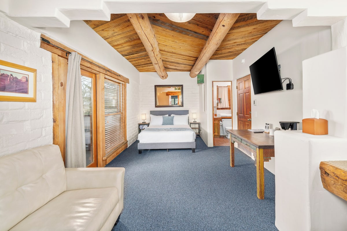 A bedroom with a bed, a wooden ceiling, a white brick wall, a TV, and a window with curtains.