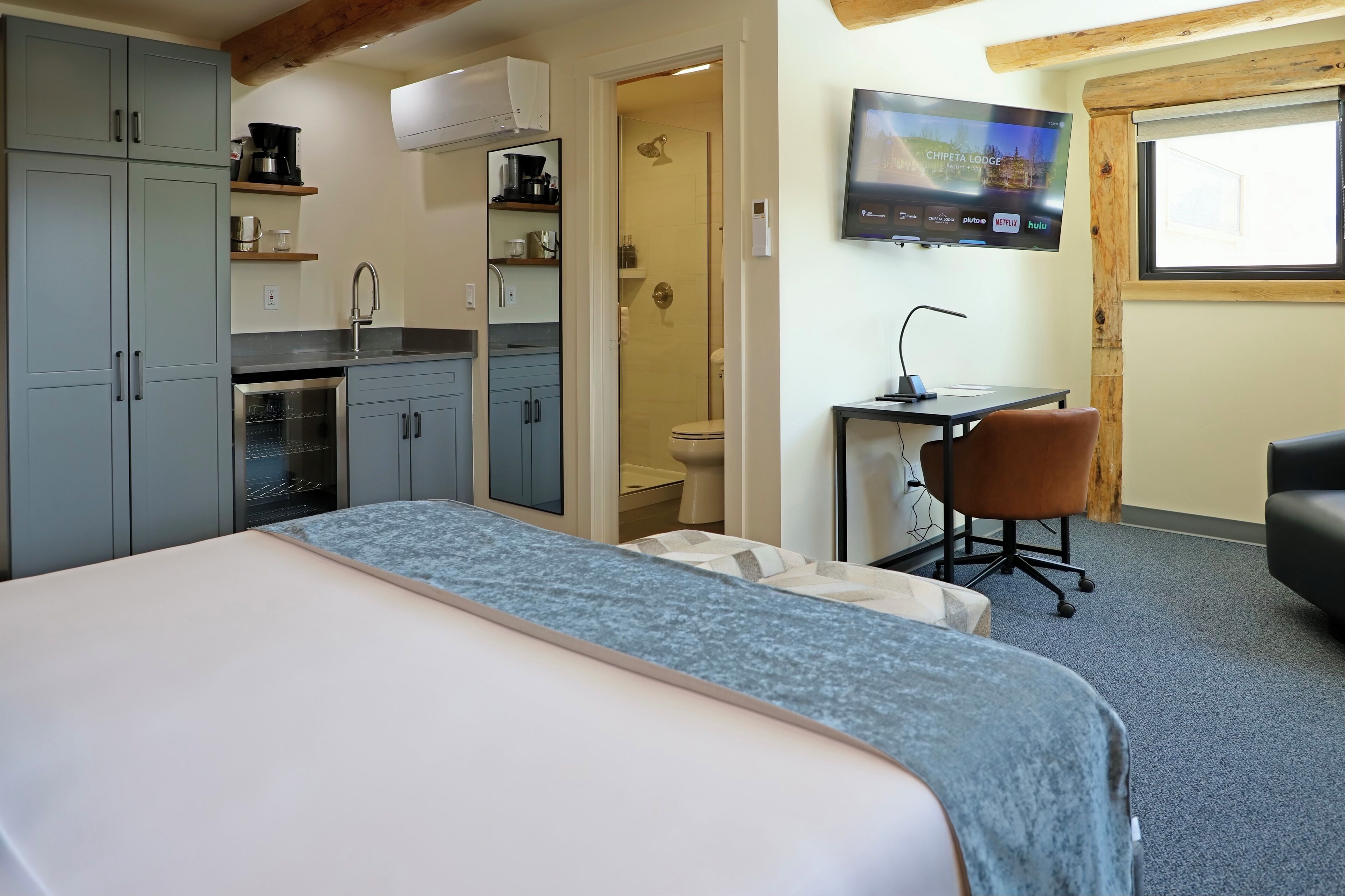 A modern hotel room with a bed, kitchenette, shower, desk, and TV.