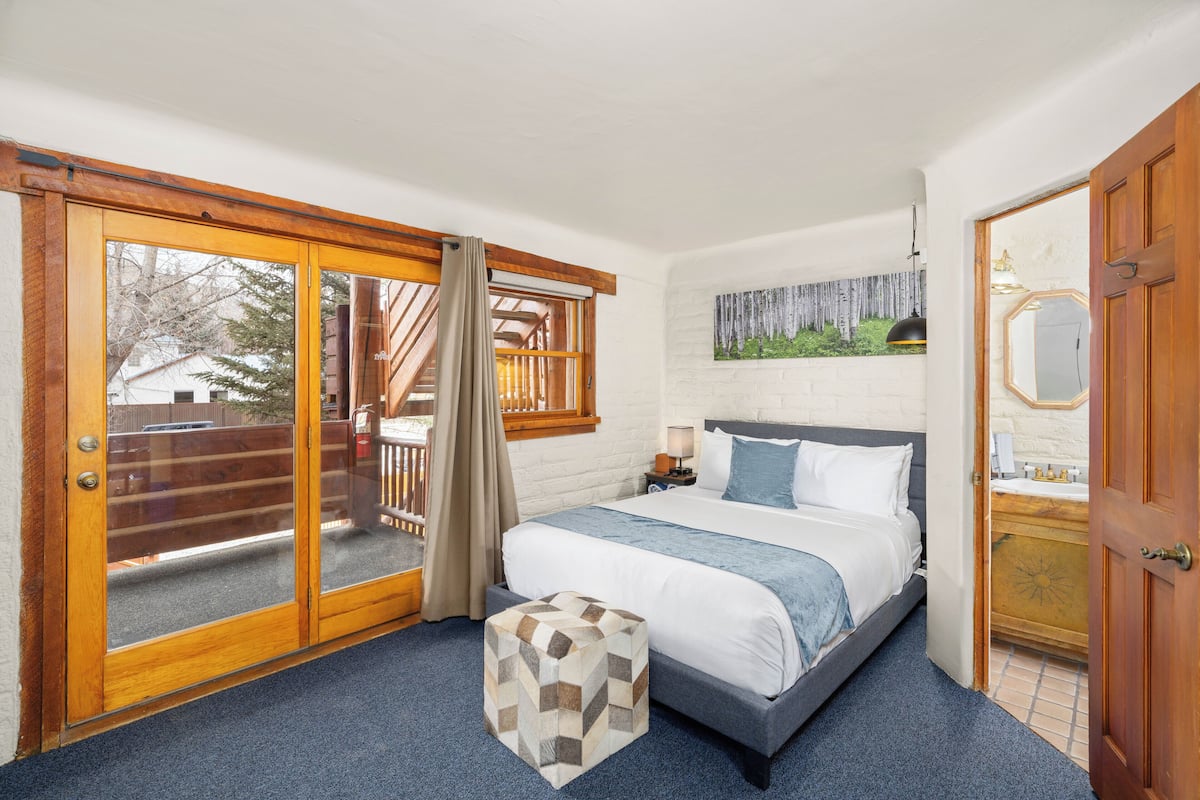A bedroom with a bed, a wooden door, a window with curtains, and a view of a deck.