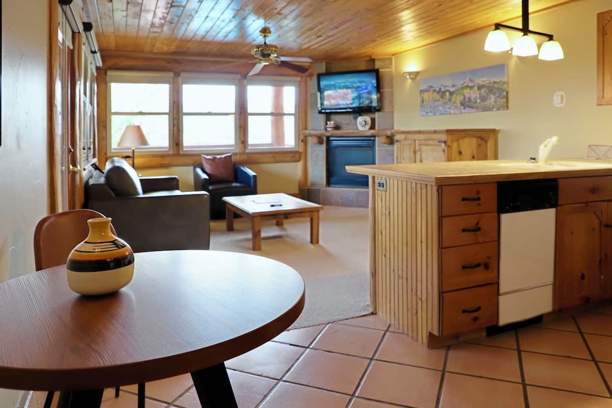A cozy living room with a sofa, a coffee table, and a wooden dining table. There is a kitchen area with a sink and cabinets.