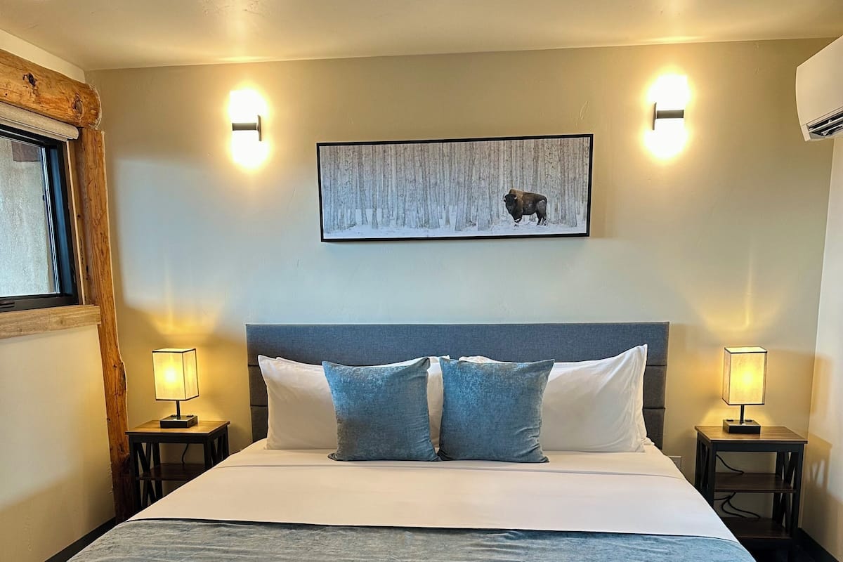 A large bed with a headboard, two bedside tables with lamps, a framed picture on the wall, and a wooden ceiling beam.
