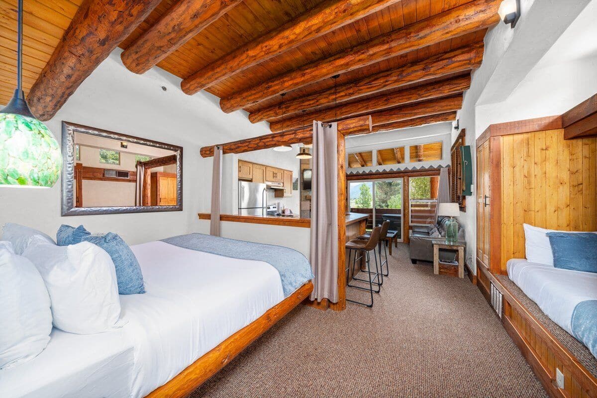 A cozy room with wooden beams, a bed, a desk, and a view of the kitchen.