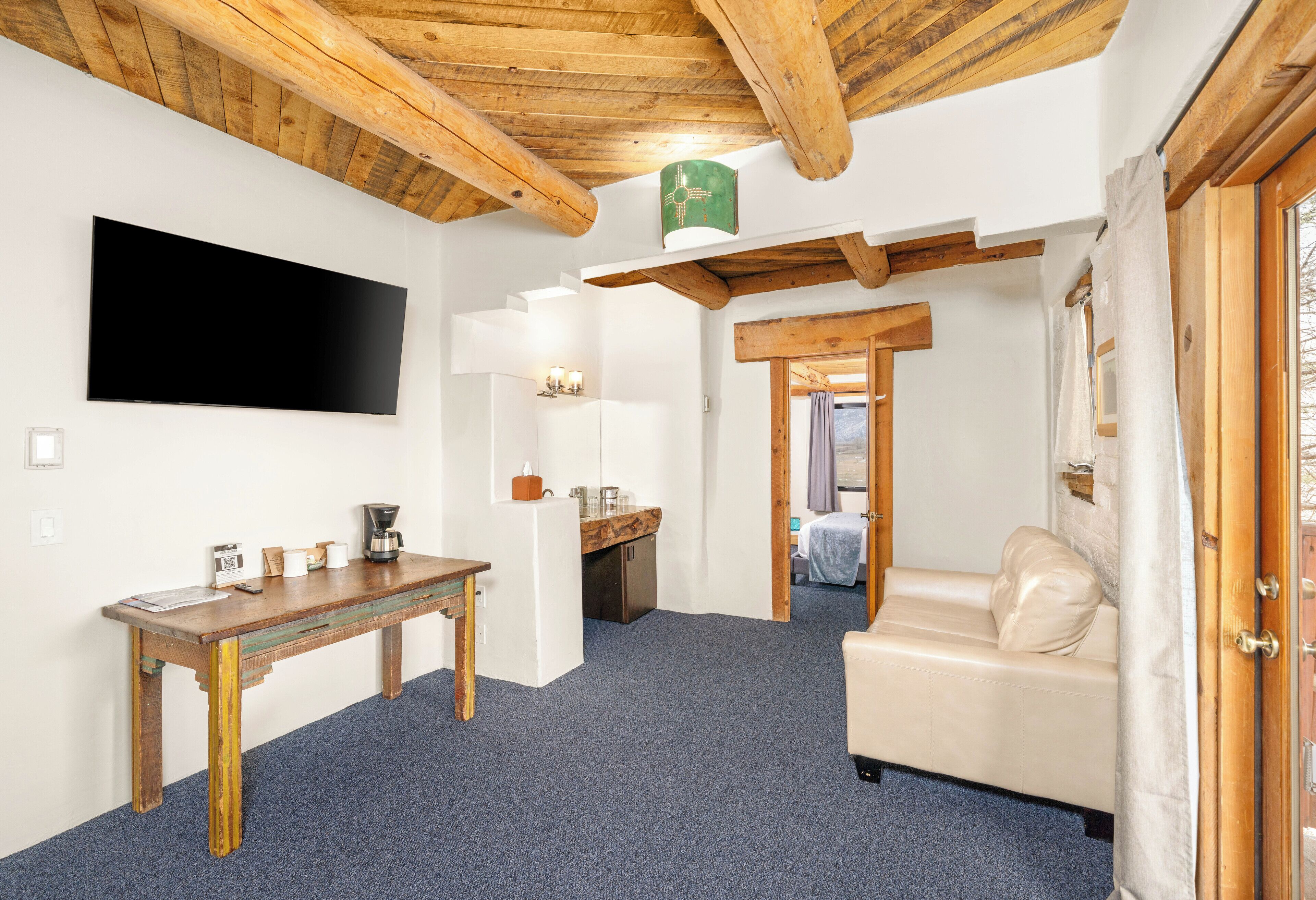 Une pièce avec un plafond en bois, un téléviseur à écran plat, une table en bois, un canapé et une salle de bain visible à travers une porte ouverte.