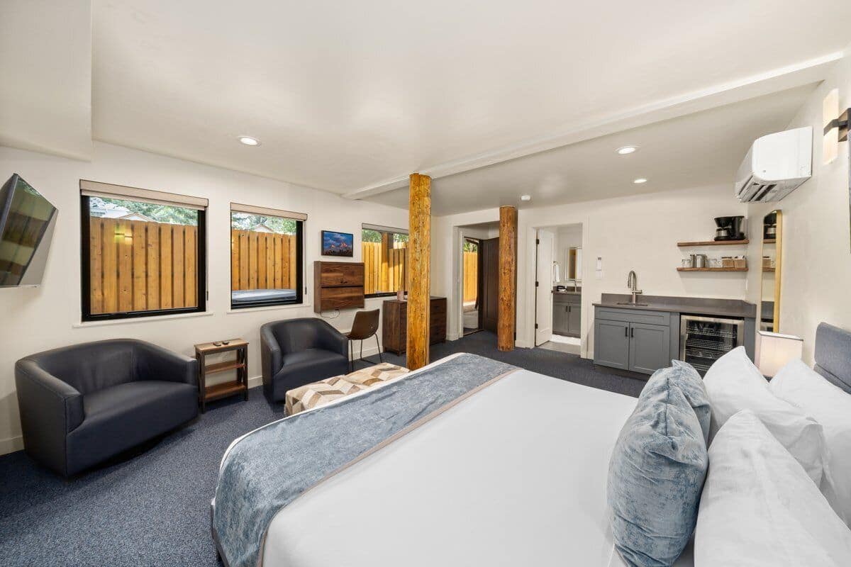 A modern hotel room with a large bed, two sofas, a small table, and a kitchenette.