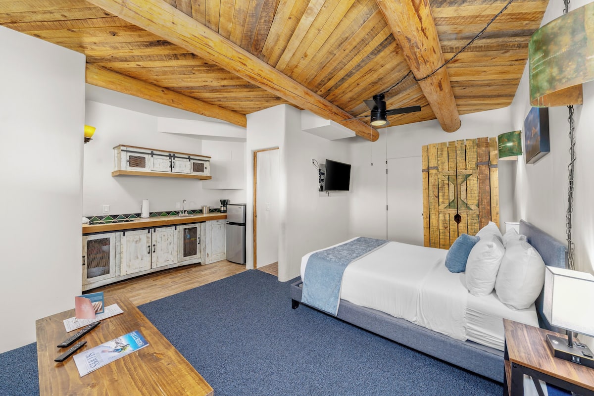 A bedroom with a wooden ceiling, a bed with white bedding, a blue carpet, a wooden table with papers, and a kitchenette with white cabinets.