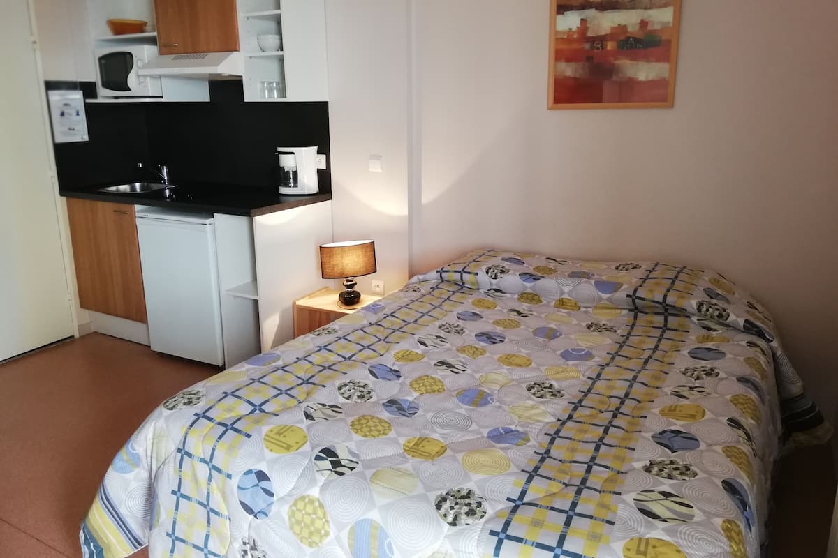 Une chambre d’hôtel compacte comprenant un coin cuisine, un lit avec une couette à motifs, une lampe de chevet et un tableau au mur.