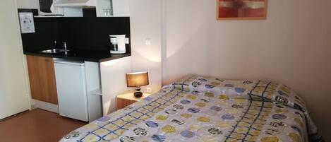 A compact hotel room with a kitchenette, a bed with a patterned duvet, a bedside lamp, and a painting on the wall.