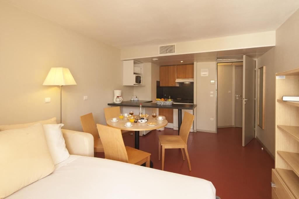 Hotellihuone, jossa on sänky, ruokapöytä tuoleineen, pieni keittiö ja lamppu.