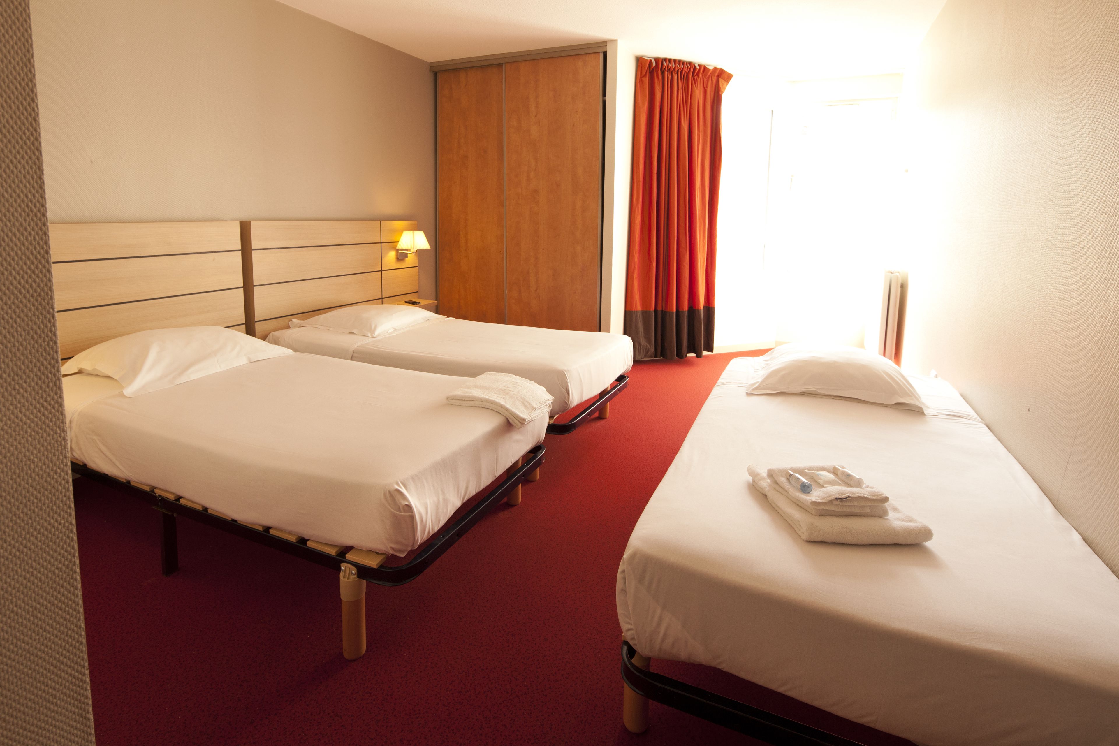 Une chambre d’hôtel avec deux lits simples, une armoire en bois, une moquette rouge et une fenêtre avec des rideaux.