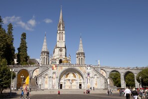 A historic cathedral with a large central arch and two spires, surrounded by a plaza with people walking. - Villa Mariale (Lourdes)