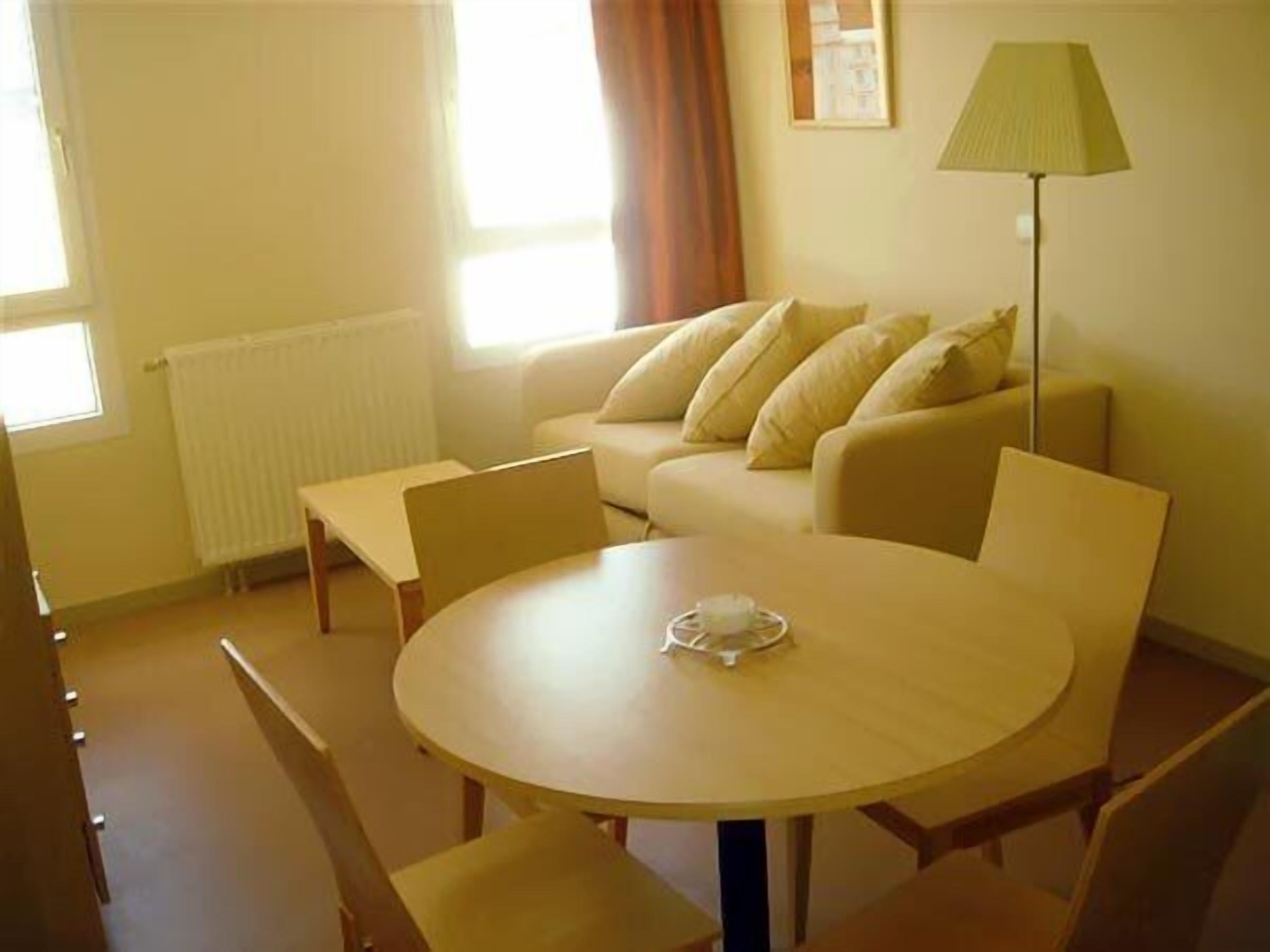 A living room with a round wooden table, two chairs, a sofa with cushions, a radiator, and a floor lamp.