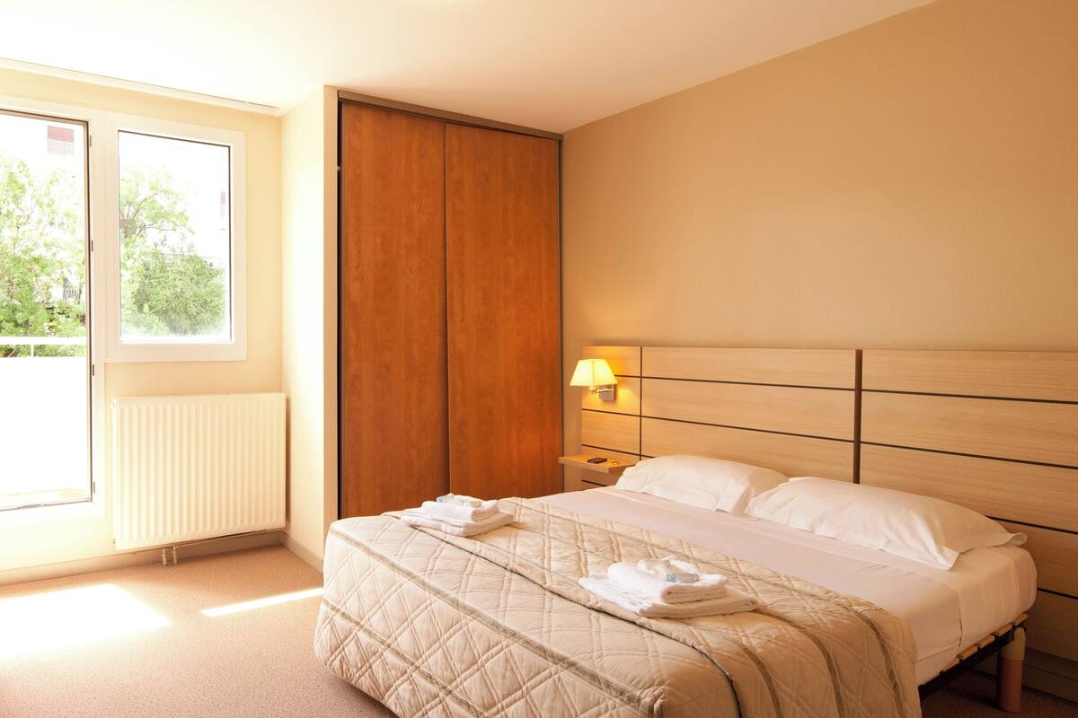 Une chambre d’hôtel avec un grand lit, une table de chevet avec une lampe, une fenêtre donnant sur un paysage verdoyant et une armoire en bois.