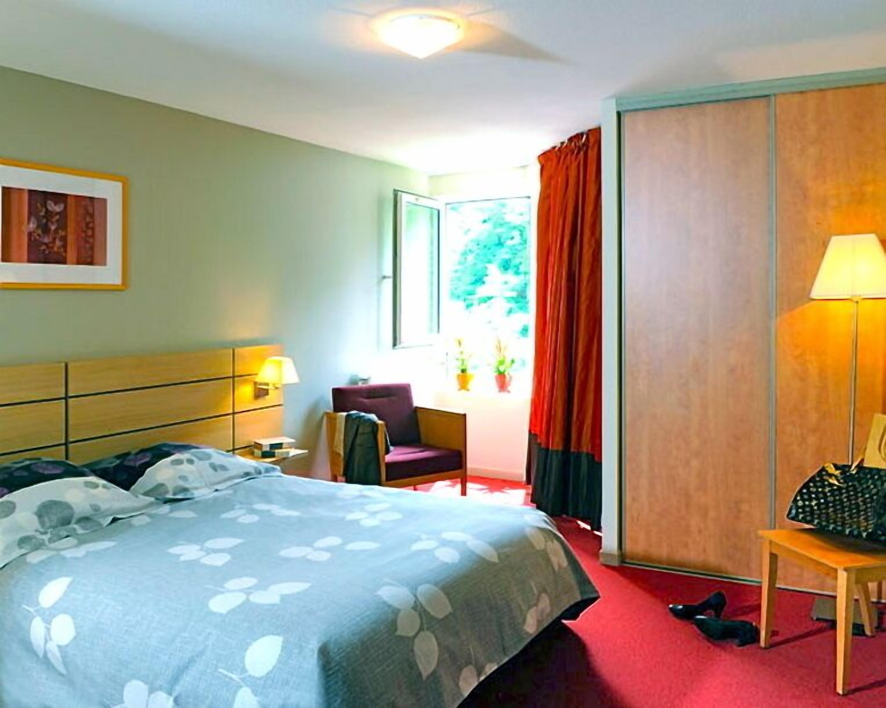A hotel room with a bed, a chair, a desk, a lamp, and a window with curtains.
