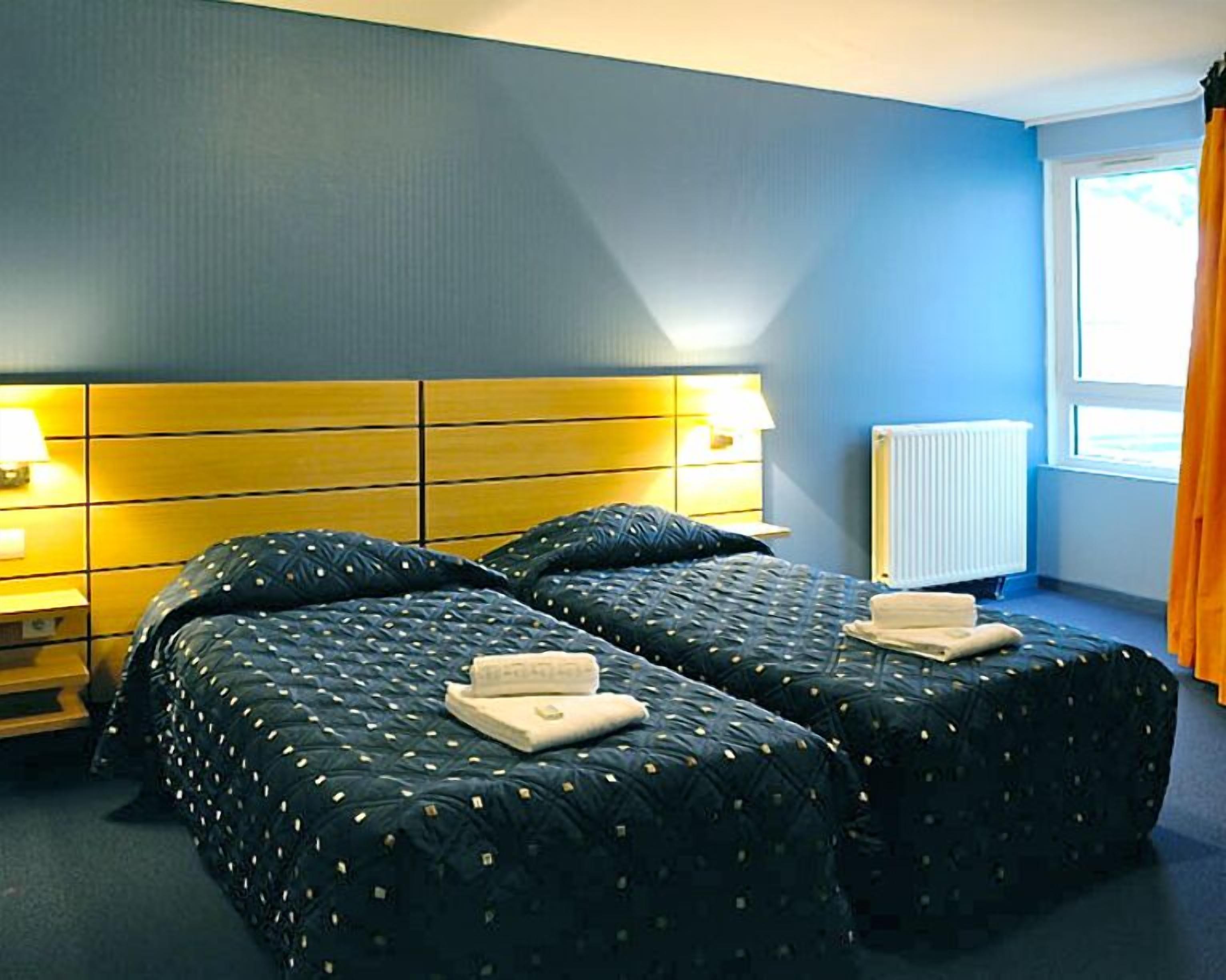 A hotel room with two beds, a wooden headboard, a radiator, and a window with curtains.