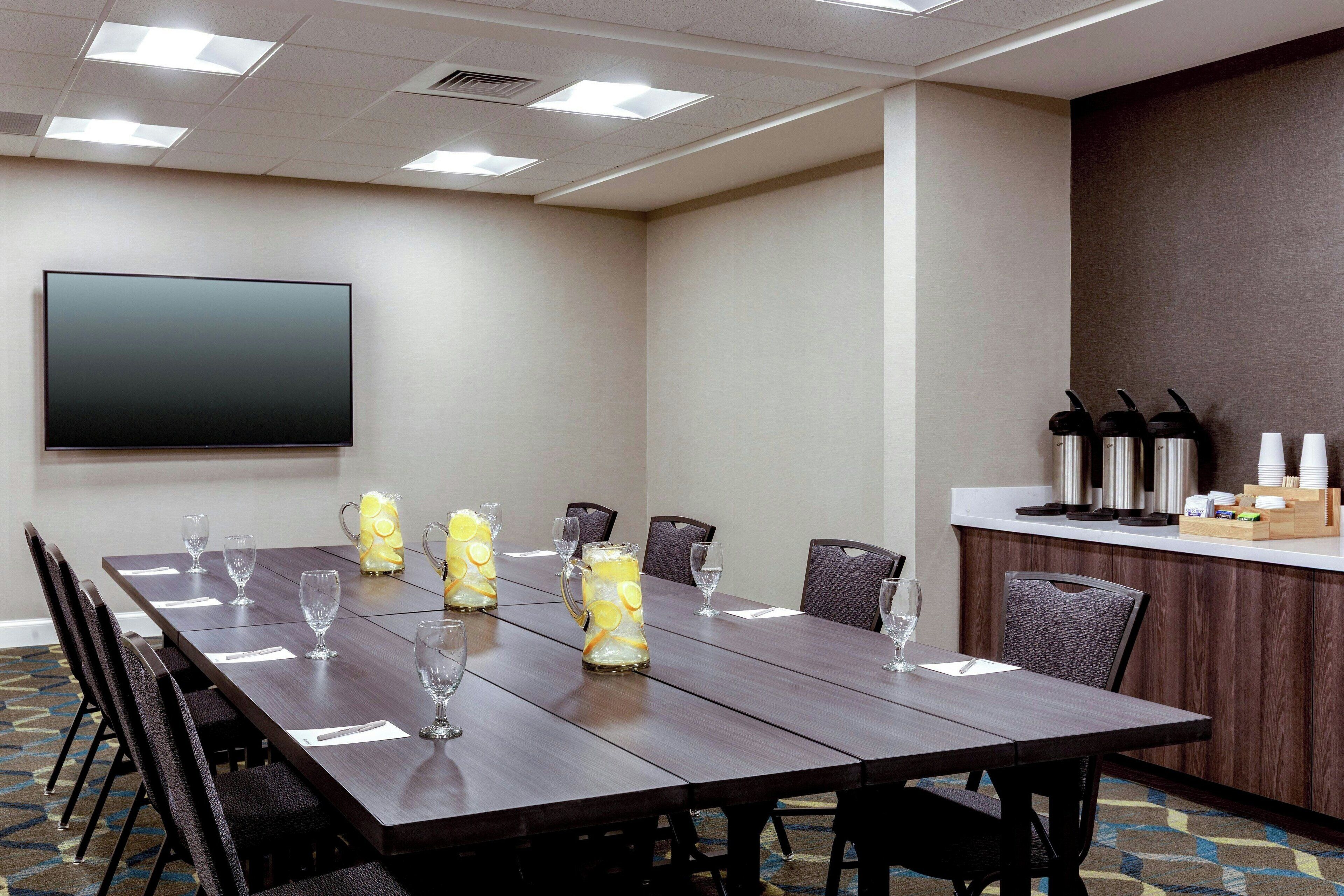 Uma sala de conferências com uma mesa comprida, cadeiras, uma TV de tela plana e um espaço de kitchenette com cafeteiras e xícaras.