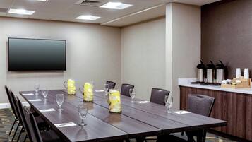 Uma sala de conferências com uma mesa comprida, cadeiras, uma TV de tela plana e um espaço de kitchenette com cafeteiras e xícaras.
