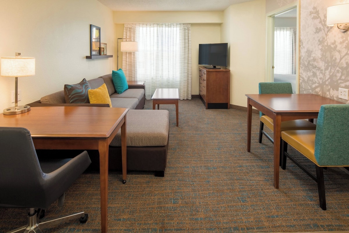Habitación de hotel con sofá, una mesita, televisor y escritorio con sillas.