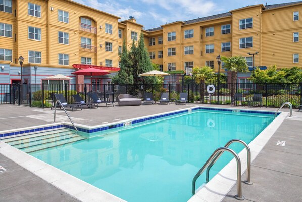 A swimming pool with a blue tiled edge, surrounded by a fence. There are lounge chairs and umbrellas, and a building with yellow walls in the background. - Residence Inn by Marriott Portland North (Portland)