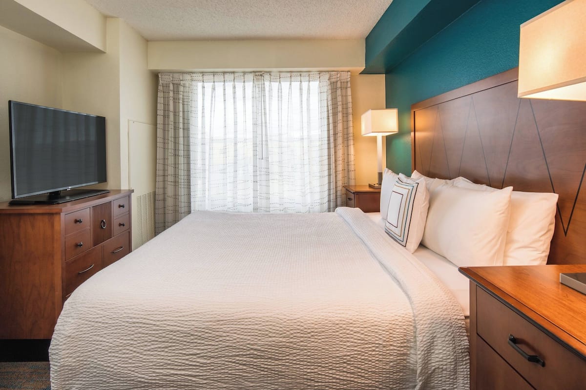 Habitación de hotel con una cama grande, un televisor sobre un tocador de madera, una mesita de noche con lámpara y una ventana con cortinas.