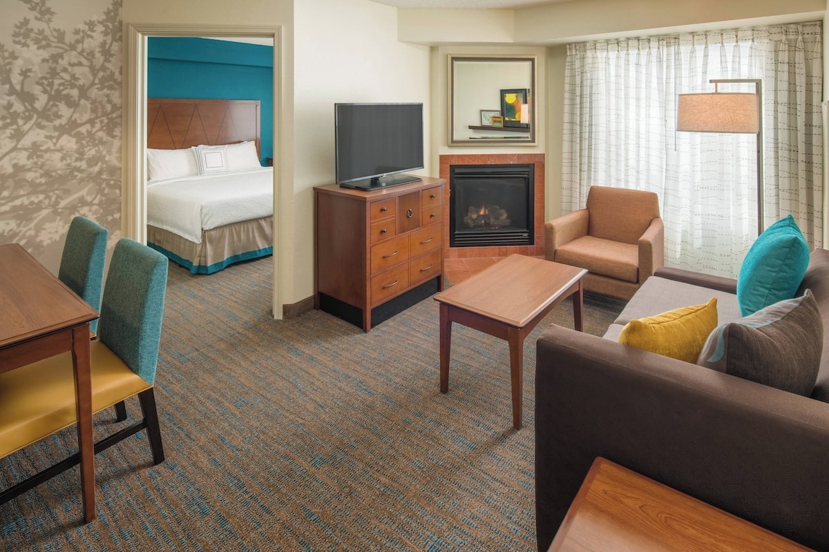 Una habitación de hotel con cama, televisión sobre un mueble de madera, chimenea, sofá, mesa de centro y una mesa de comedor con sillas.