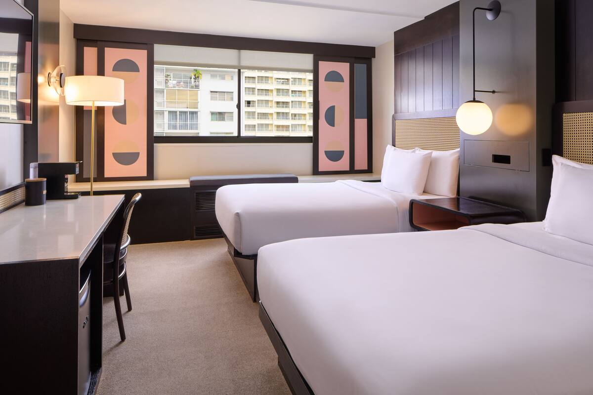 Une chambre d’hôtel avec deux lits, un bureau, une chaise, une lampe et une vue sur les immeubles par la fenêtre.