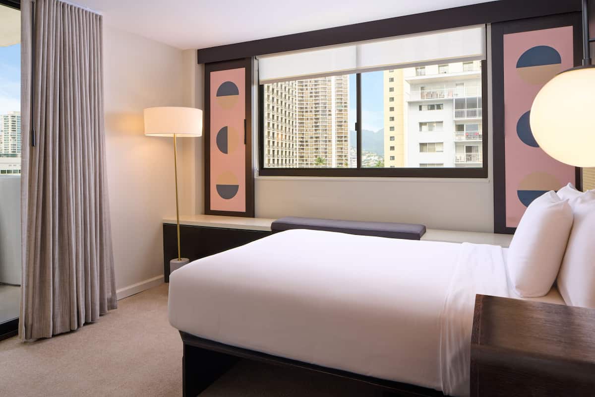 Une chambre d’hôtel avec un lit, une fenêtre donnant sur un immeuble, une lampe et un rideau.