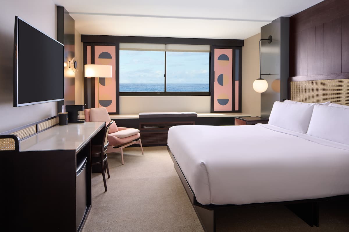 Une chambre d’hôtel équipée d’un lit, d’un bureau, d’une chaise et d’une grande fenêtre donnant sur la mer.