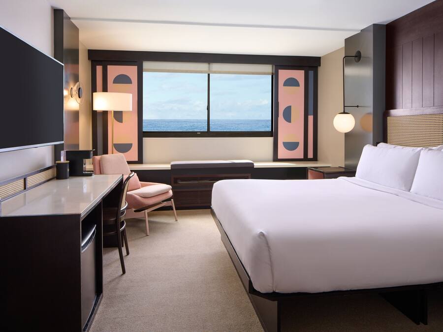 A hotel room with a bed, desk, chair, and a large window with a view of the sea.