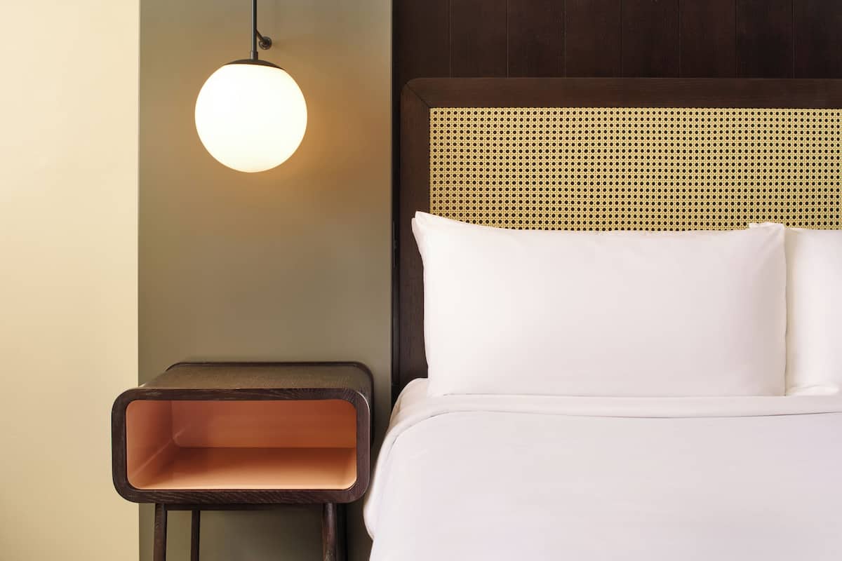Un lit bien fait, avec du linge de lit blanc, un luminaire rond fixé au mur et une petite table de chevet en bois, avec un plateau marron et un intérieur orange.