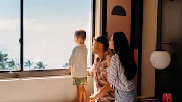 A family of three by a large window with a view of palm trees and the ocean.