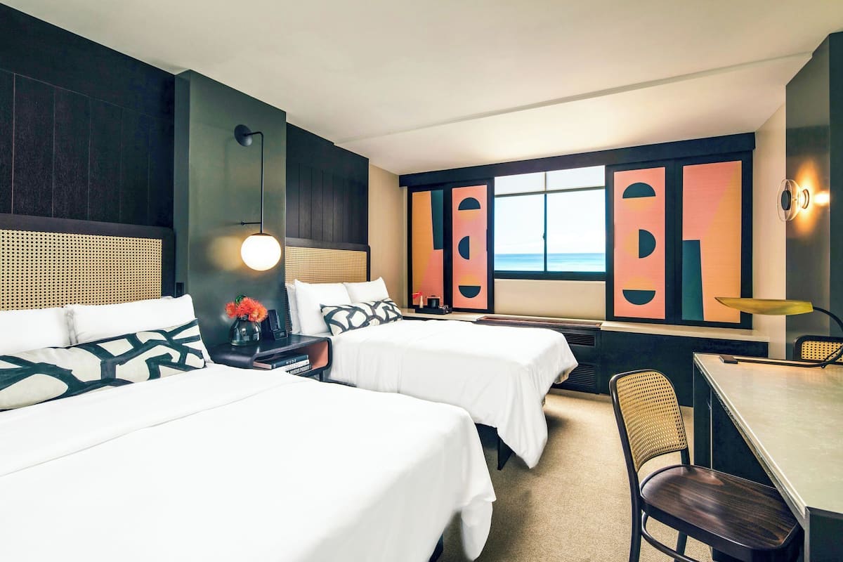 Habitación de hotel con dos camas, un escritorio, una silla, una lámpara y un ventanal con vistas.