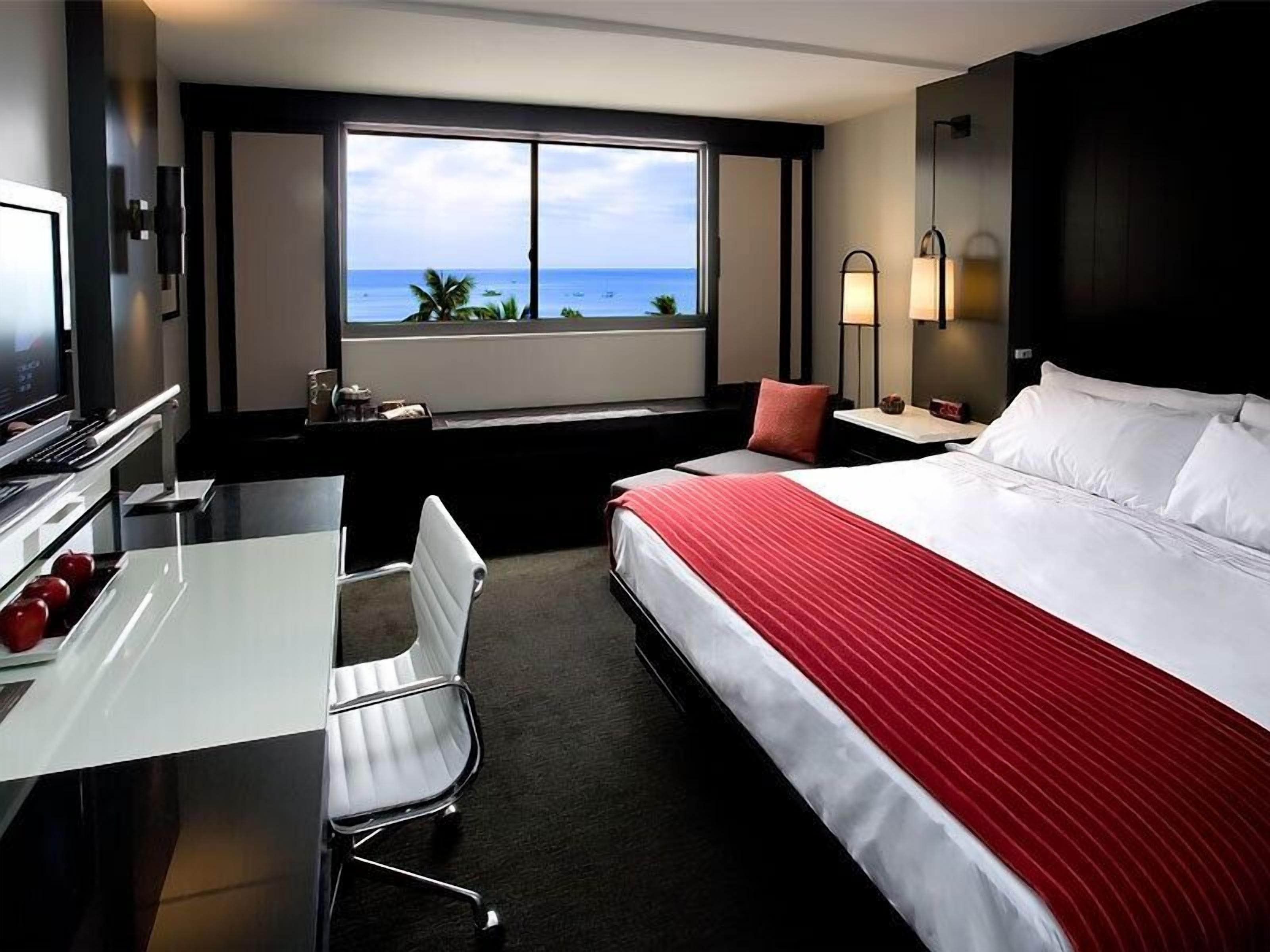 Une chambre d’hôtel comprenant un lit, un bureau, une chaise, une télévision, une fenêtre donnant sur l’océan et une table de chevet avec une lampe.