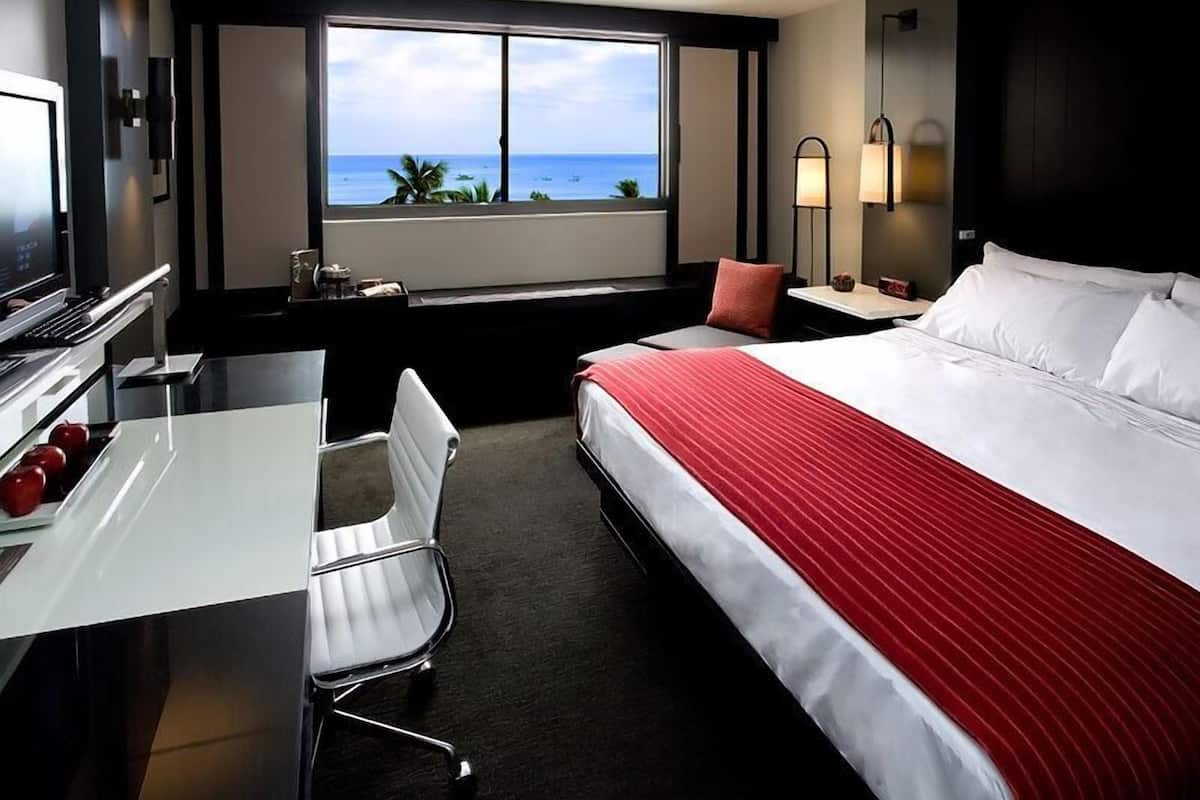 Habitación de hotel con cama, escritorio, silla, televisor, ventana con vista al océano y mesita de noche con lámpara.